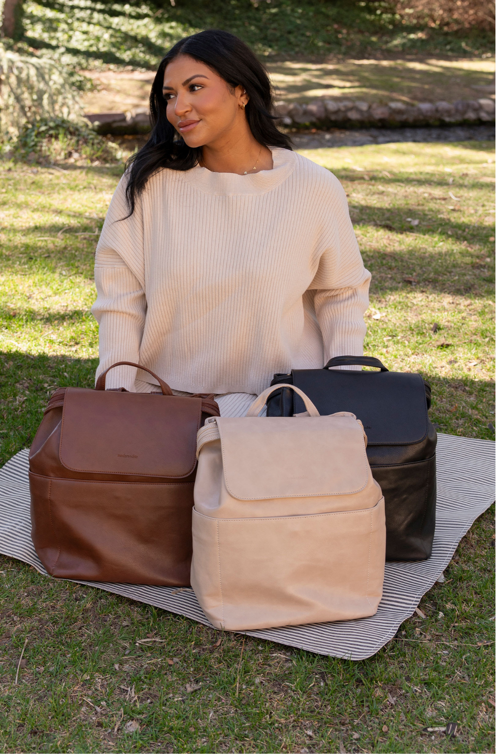 A woman kneeling on a black and white striped blanket sitting on the grass with three different Modern+Chic Brielle 2.0 Vegan Leather Convertible Backpack Purses for Women sitting in front of her in the shades dark brown, taupe and black.