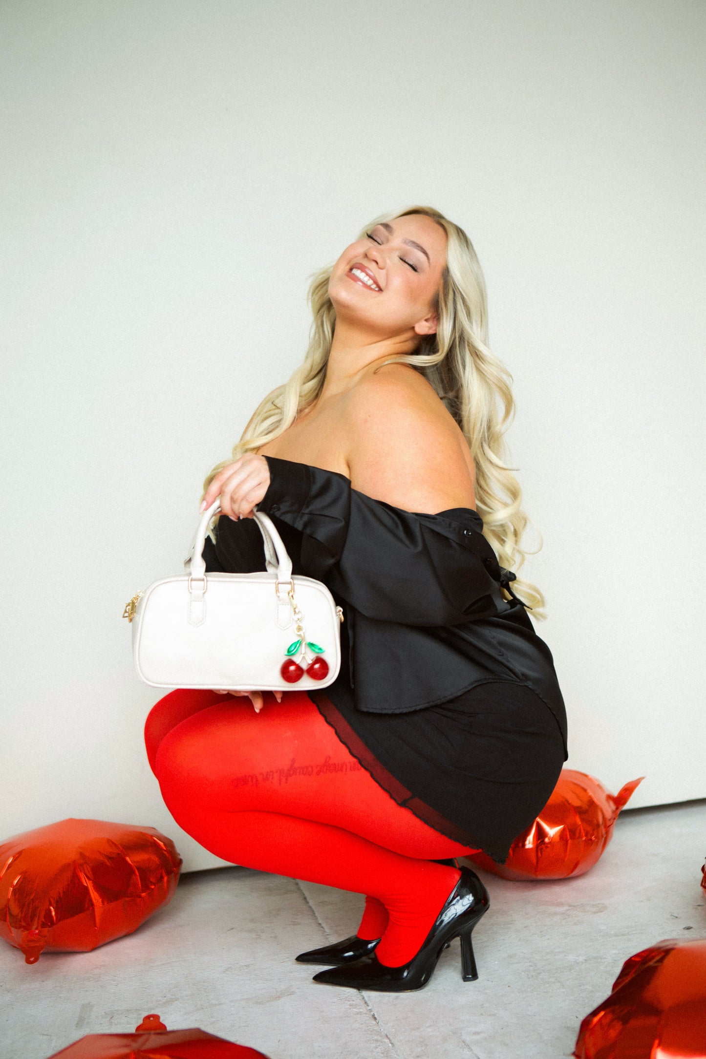 A woman wearing a black stress squatting down holding her cream Modern + Chic Remi Vegan Leather handbag with and a large cherry charm hanging from the strap.