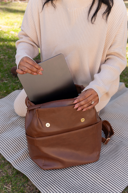 A woman kneeling on a black and white striped putting a laptop into a dark brown Modern+Chic Brielle 2.0 Vegan Leather Convertible Backpack Purse for Women with gold hardware. 