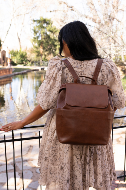 An image of a dark-haired woman facing away wearing a cream and brown floral dress with a dark brown Modern+Chic Brielle 2.0 Vegan Leather Convertible Backpack Purse for Women. 