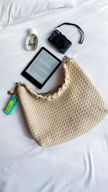 An ivory Modern+Chic Kelly Woven Convertible Crossbody Shoulder Bag with a variety of items including perfume, a camera and a kindle.
