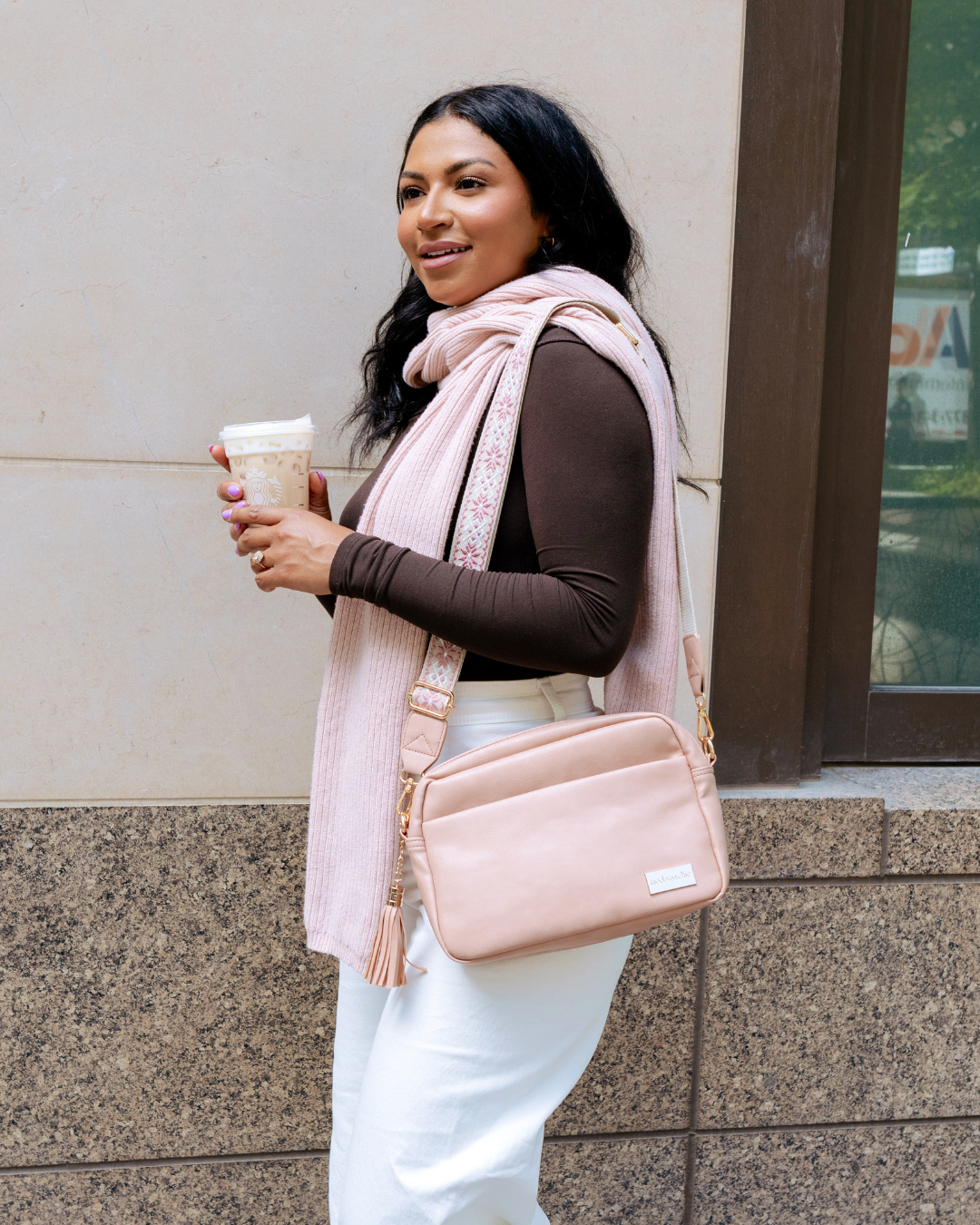 Woman holding a coffee cup, wearing a pink scarf and carrying a dusty blush Modern + Chic Alice Crossbody Vegan Leather bag  against a building background.