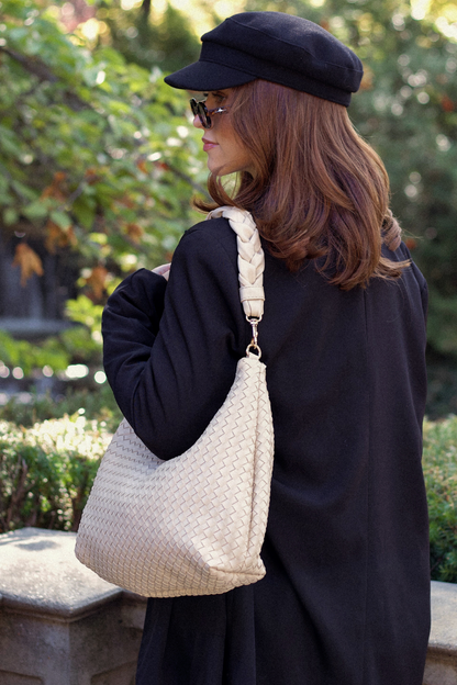 Woman in black coat and cap holding an ivory Modern+Chic Kelly Woven Convertible Crossbody Shoulder Bag outdoors.
