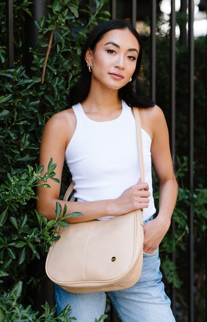 A woman standing in front of greenery and a fence wearing a cream Modern + Chic Marissa crossbody vegan leather handbag. 