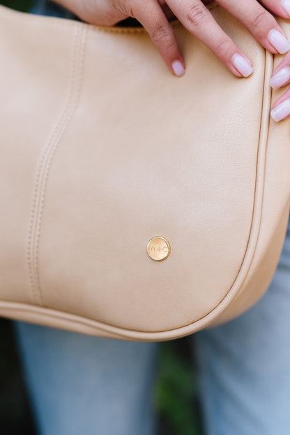 A close-up shot of a circular gold logo with m+c etched into it on a dusty blush Modern + Chic Marissa Vegan Leather Crossbody Handbag. 