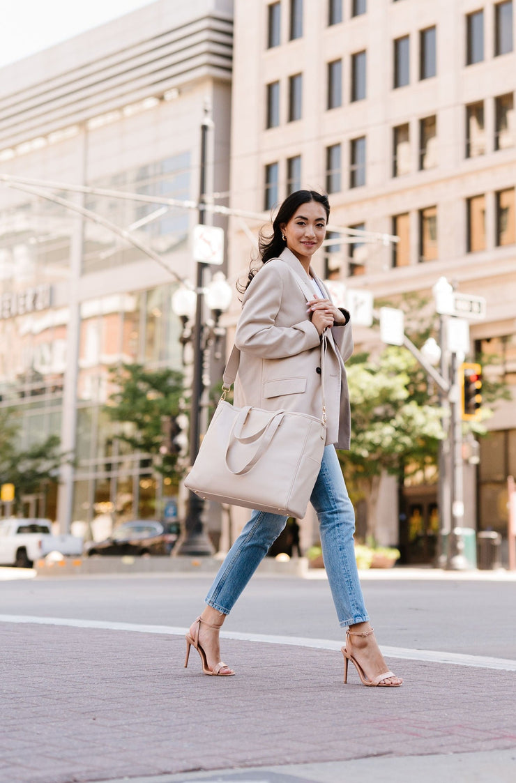 A woman crossing a city street wearing a cream tote on her shoulder.