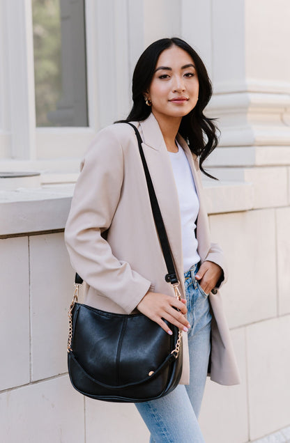 A woman standing in front of a white building wearing a black Modern + Chic Marissa vegan leather crossbody bag over shoulder.