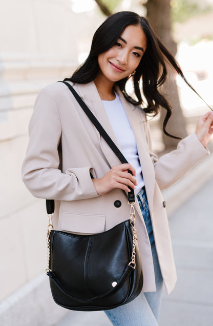 A woman standing in front of a white building wearing a black Modern + Chic Marissa vegan leather crossbody handbag.