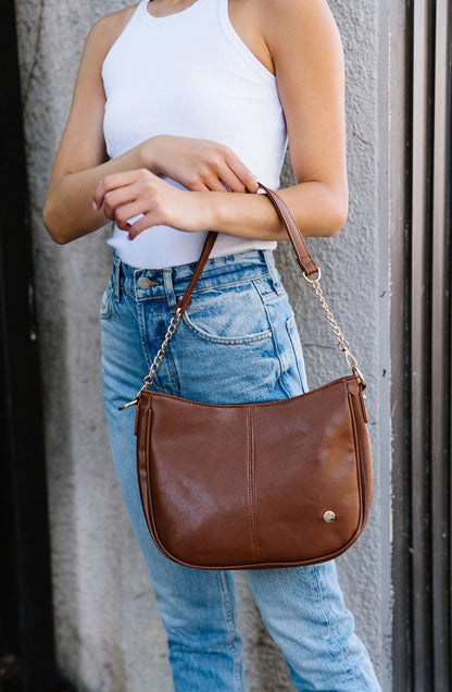 A woman wearing a brown Modern + Chic Marissa Vegan Leather Saddle Crossbody Bag handbag on her arm.