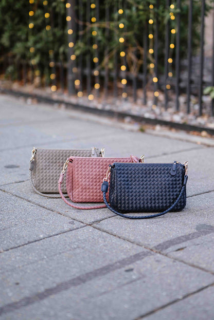 Three woven Modern + Chic Elle Woven Crossbody Bag  in gray, pink, and blue on a paved surface with blurred lights in the background.
