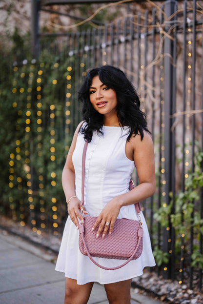 Woman in a white dress holding a pinkA woman wearing a pink Modern + Chic Elle Woven Crossbody Bag for Women crossbody against a decorative fence background.