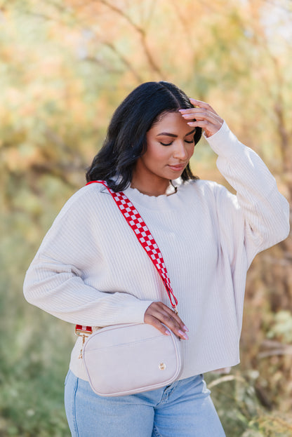 A woman wearing a light pink sweater and blue jeans is shown with a red and pink Modern+Chic Lizzie checkered adjustable strap attached to a light pink purse.