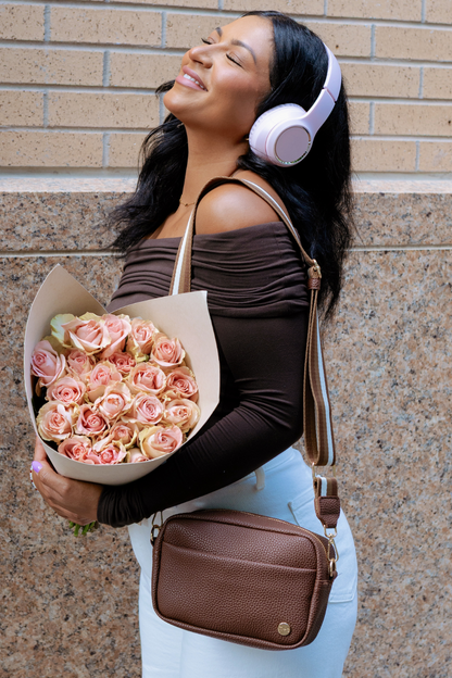 Woman wearing a brown Modern+Chic Willow Vegan Leather Convertible Crossbody Bag against a brick wall. 