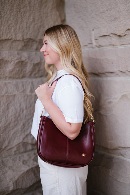 A woman wearing a wine Modern + Chic Marissa vegan leather handbag with gold chain accents on the strap over her shoulder. 