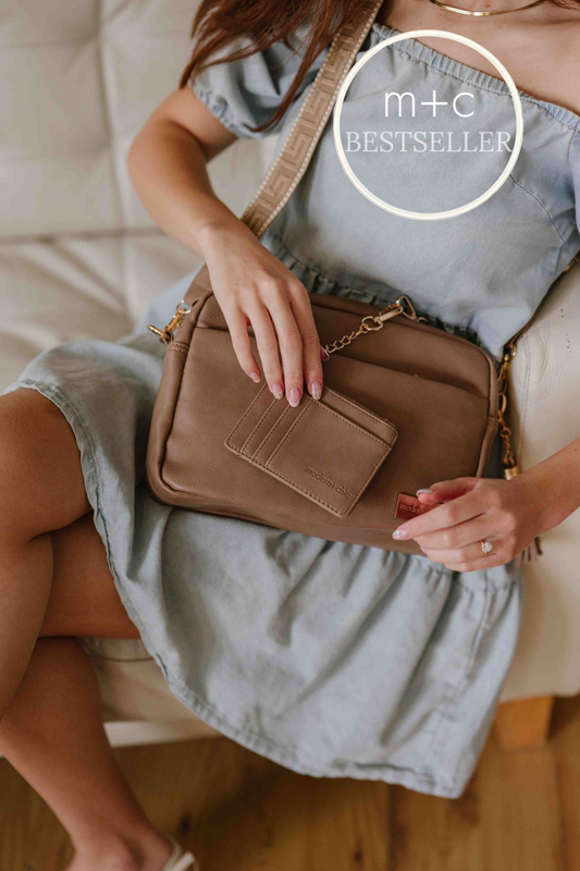 Woman holding a fawn colored Modern + Chic Alice Crossbody Vegan Leather bag, with the matching wallet and included key leash in hand with 'm+c BESTSELLER' label at the top right of the image. 