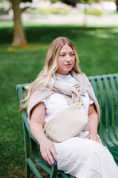 Woman sitting on a bench wearing a white t-shirt and a tan sweater tied around her shoulders and a cream criss cross style Modern + Chic Rory Oversized Belt Bag for Women with a gold zipper and hardware crossbody.