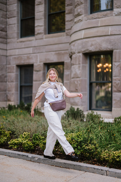 Woman wearing a white t-shirt, a sweatshirt tied around her shoulders and a brown criss cross style Modern + Chic Rory Oversized Belt Bag for Women with a gold zipper and hardware crossbody.