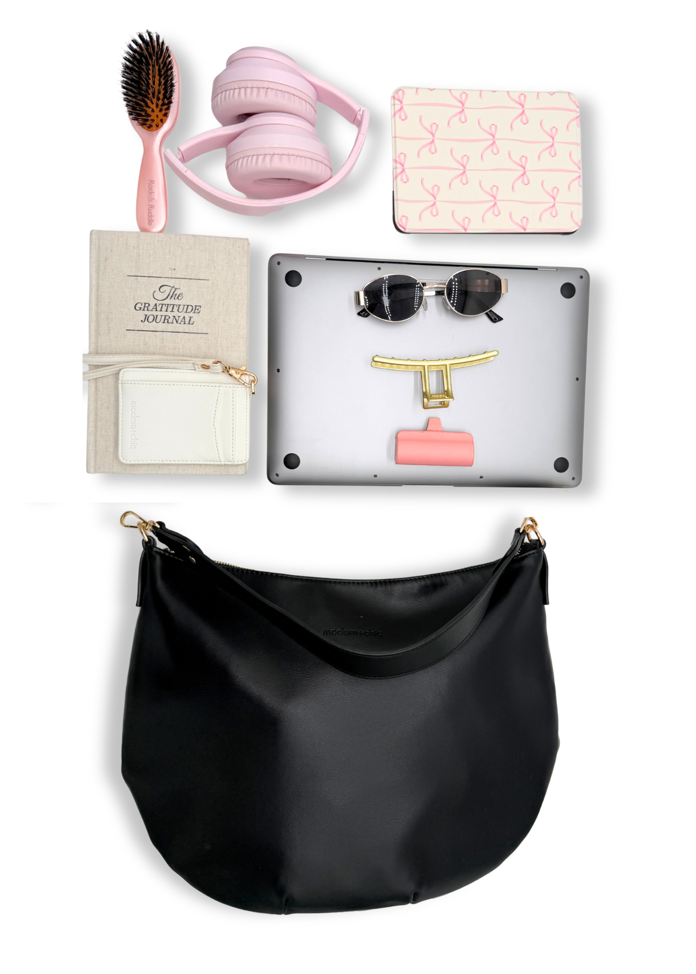 A black vegan leather Modern+Chic Savannah Slouchy Tote on a white background with pink accessories including headphones, a hairbrush, and a small notebook on a white background.