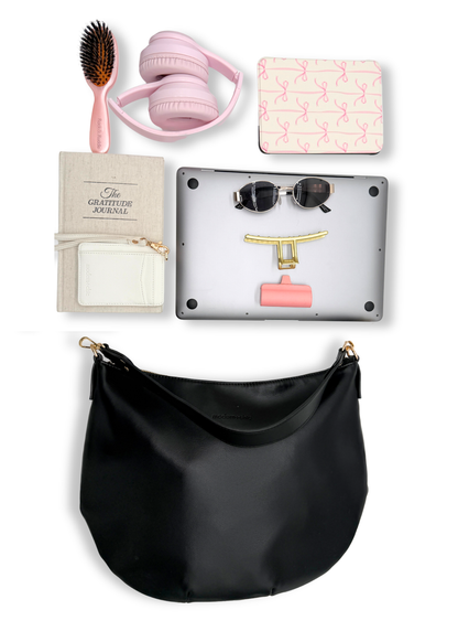 A black vegan leather Modern+Chic Savannah Slouchy Tote on a white background with pink accessories including headphones, a hairbrush, and a small notebook on a white background.