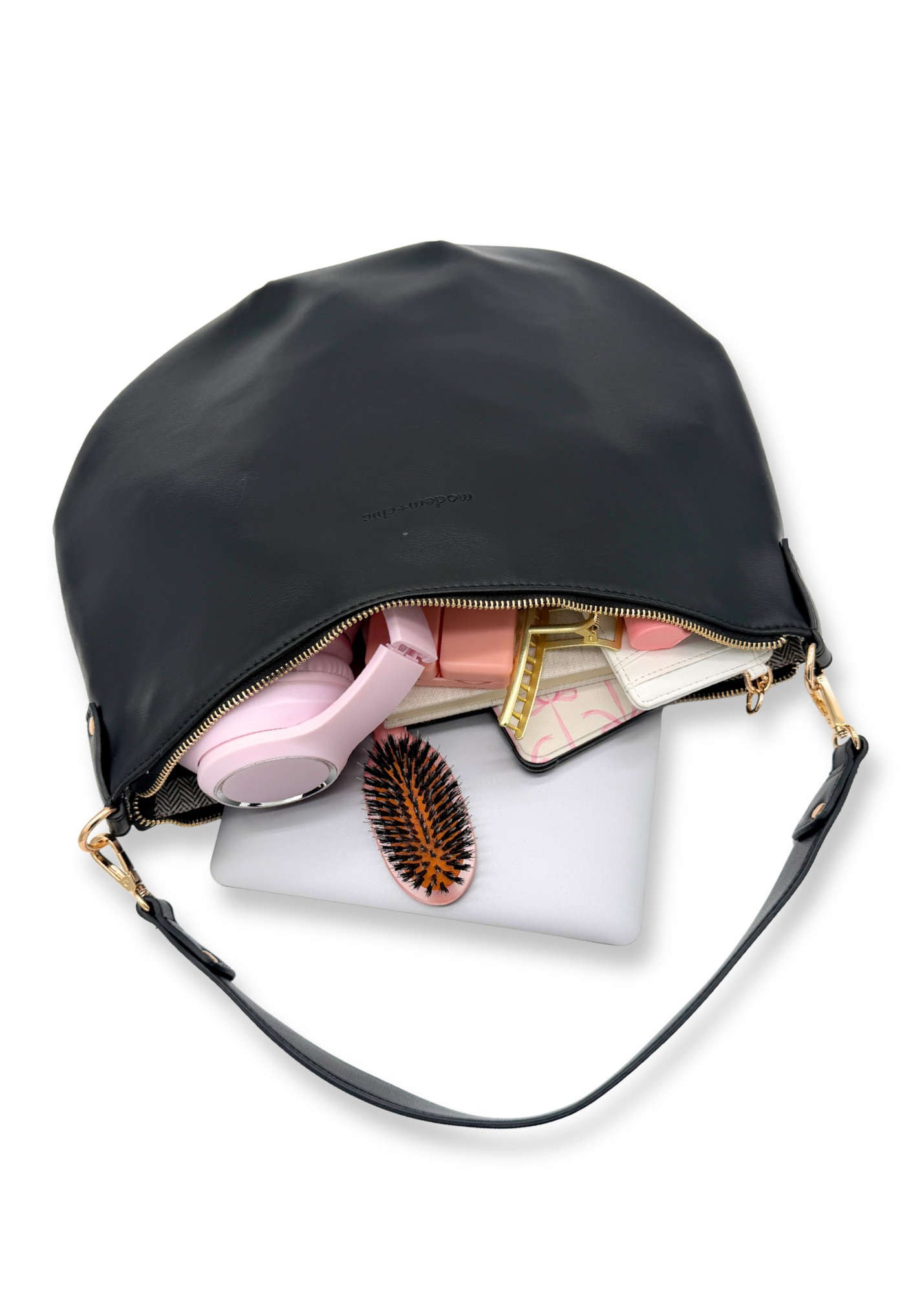 A black vegan leather Modern+Chic Savannah Slouchy Tote on a white background with a laptop, pink headphones, kindle and hairbrush on a white background. 
