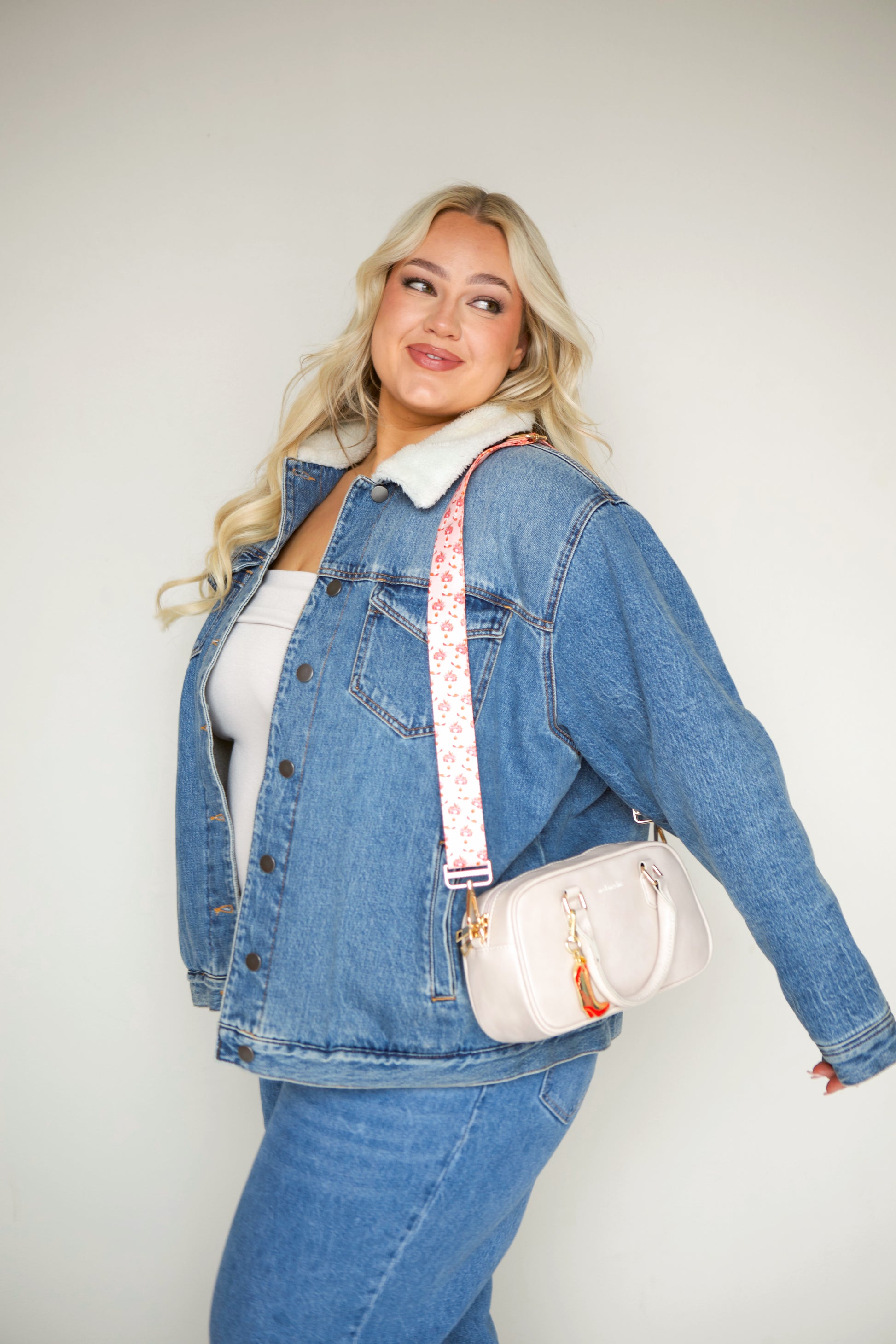 Woman wearing a denim jacket with a cream Modern + Chic Remi Vegan Leather Crossbody Purse with gold hardware, cream printed strip and charm. 