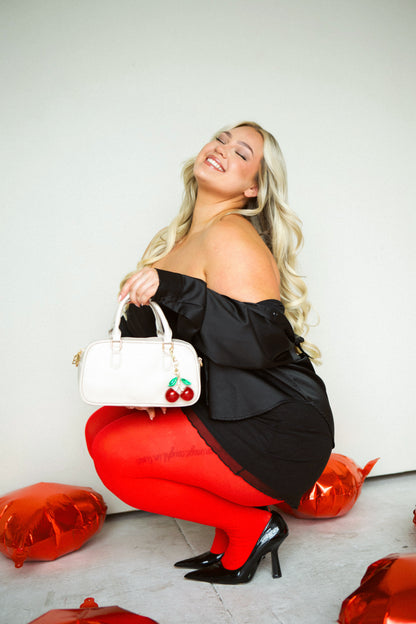  A woman wearing a black stress squatting down holding her cream Modern + Chic Remi Vegan Leather handbag with  and a large cherry charm hanging from the strap.
